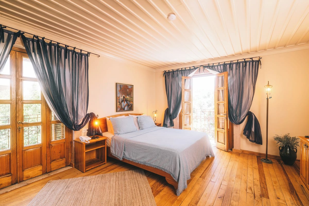 Un dormitorio con cama de madera, una mesita de noche con lámpara, un ventanal con cortinas y una planta en maceta.
