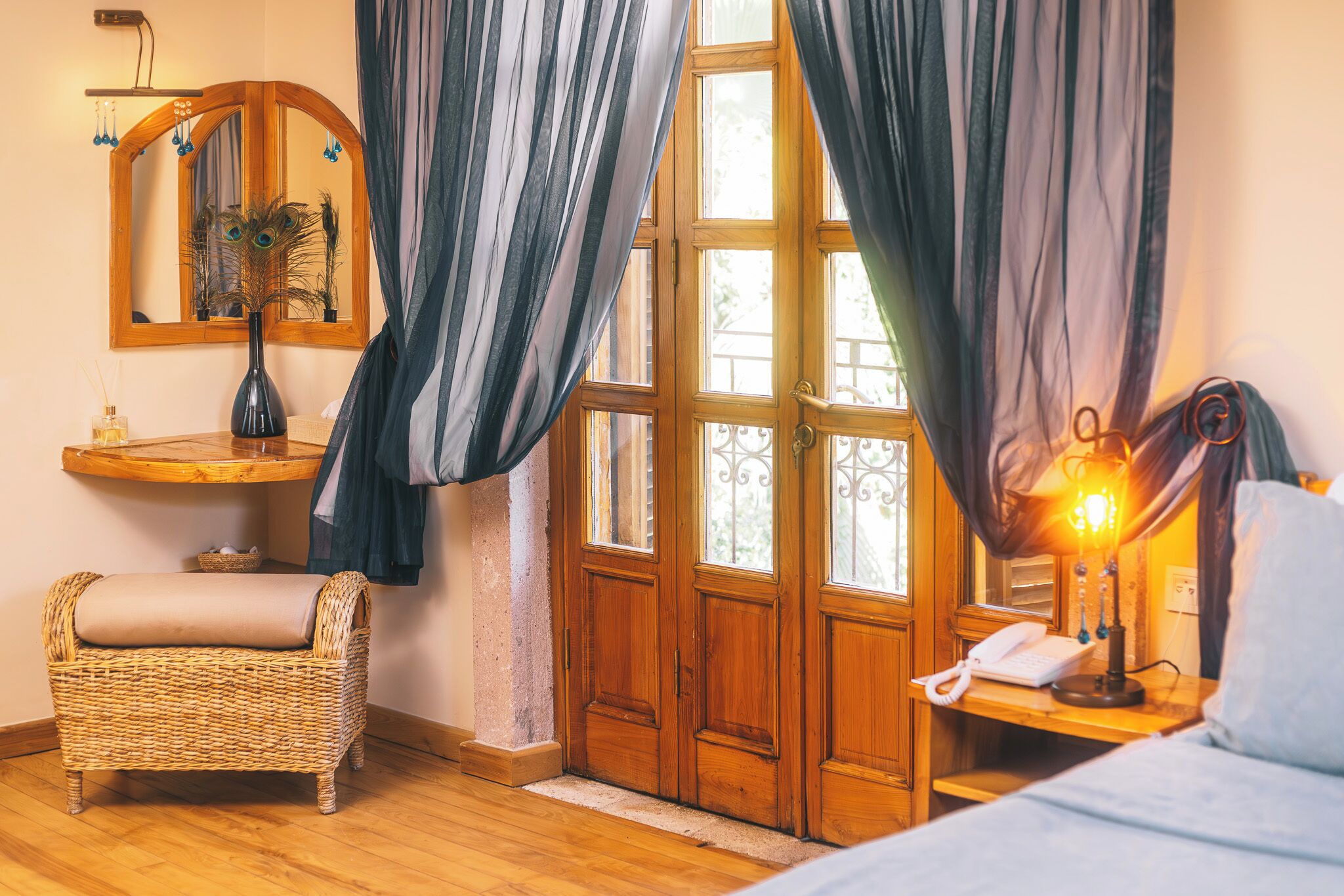 Ein Zimmer mit Holztür, einem Korbsessel, einem Nachttisch mit Lampe und einem Spiegel an der Wand.
