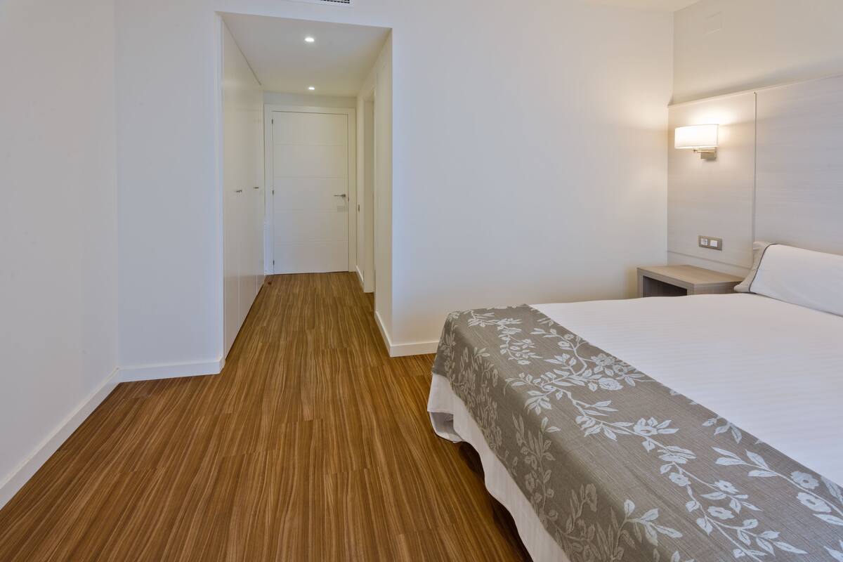 A hotel room with a wooden floor, a bed with a floral bedspread, a nightstand, and a wall-mounted light.
