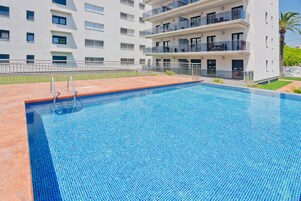 A swimming pool with a tiled edge, surrounded by a white building with balconies and a metal fence.