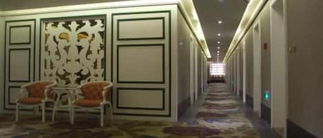 A hotel corridor with a patterned carpet, a seating area with chairs, and a decorative wall panel.