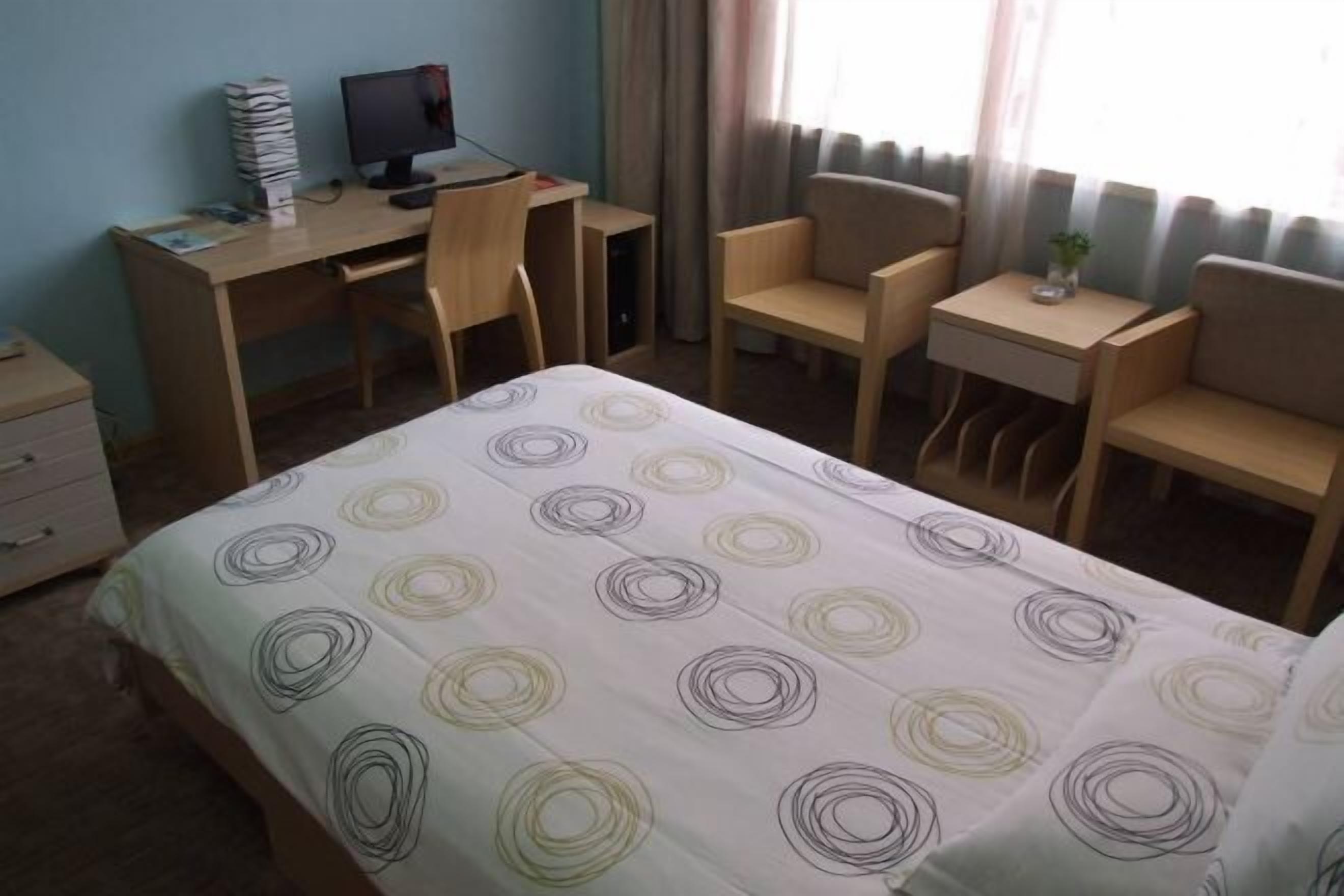 A hotel room with a bed, a desk with a computer, two chairs, a small table, and a nightstand.
