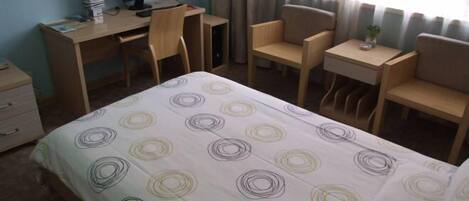 A hotel room with a bed, a desk with a computer, two chairs, a small table, and a nightstand.