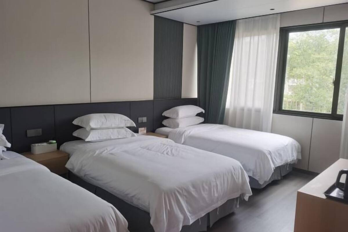 A hotel room with two single beds, a nightstand with a lamp, a desk with a chair, and a window with curtains.