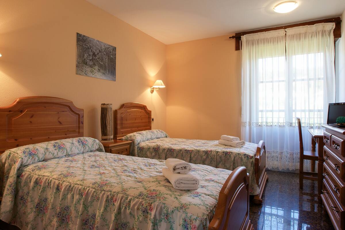 A hotel room with two wooden beds, a desk with a chair, and a window with curtains.