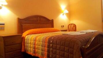 Una cama bien hecha con colcha, una mesita de noche de madera, una silla y dos lámparas fijadas a la pared.