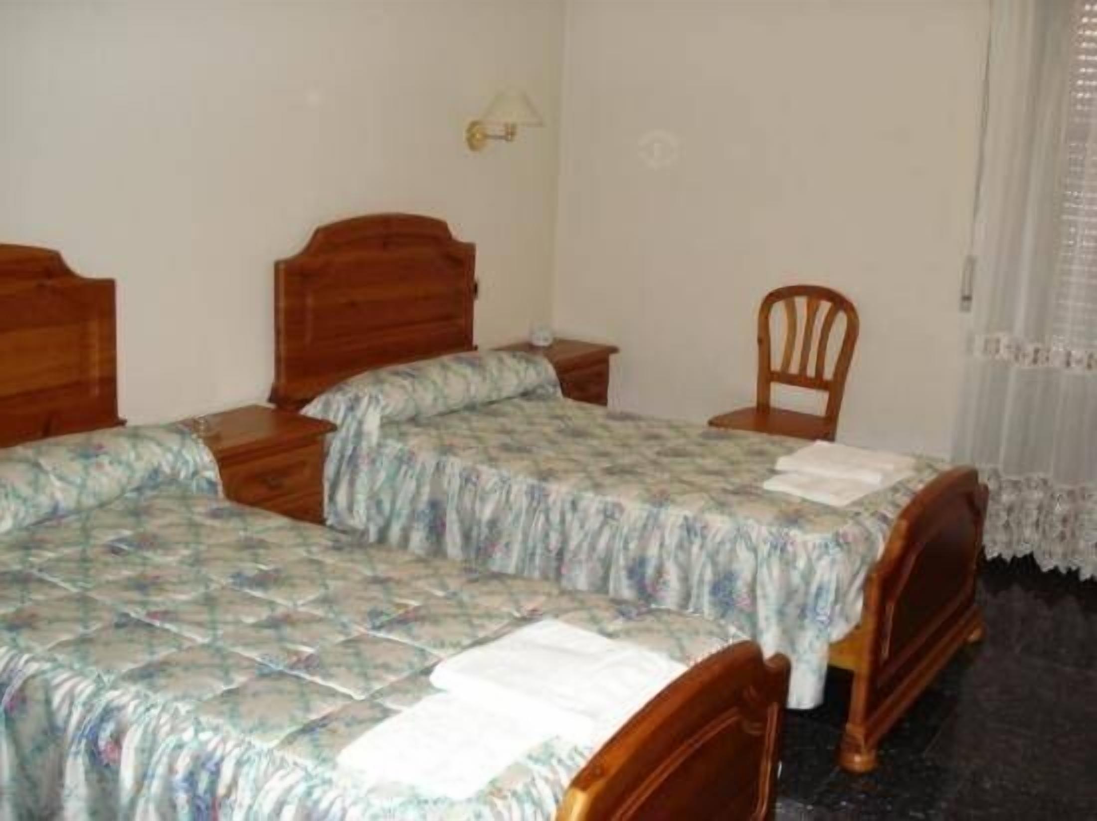 A simple room with two wooden beds, a small table, a chair, and a lamp.