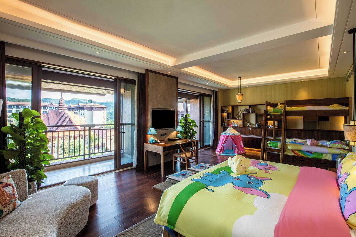 Ein Hotelzimmer mit Balkon, Schreibtisch mit Fernseher, Bett mit farbenfroher Elefantendecke und einem kleinen pinken Zelt.