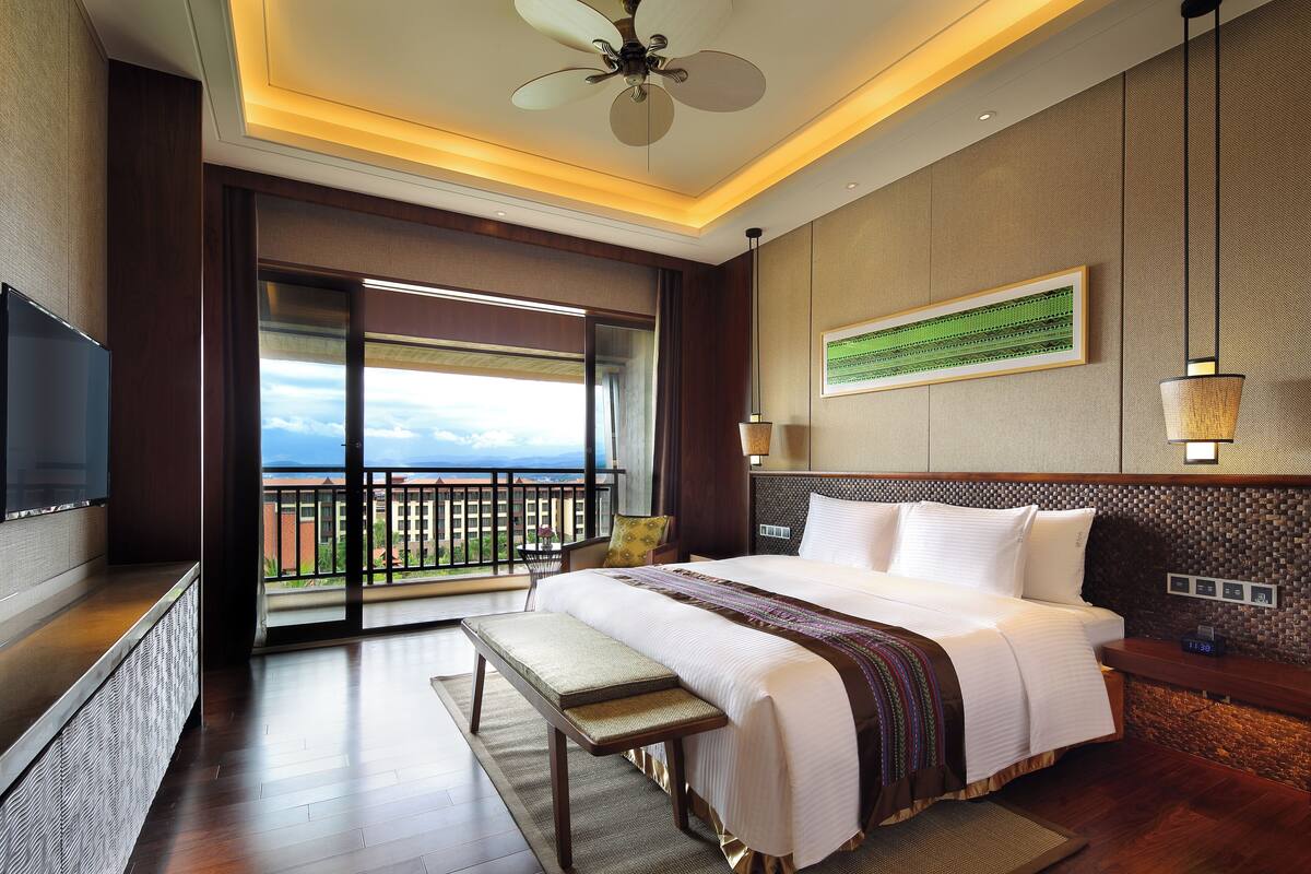 A hotel room with a large bed, a bench, a TV, a ceiling fan, a balcony with a view, and a wall-mounted artwork.