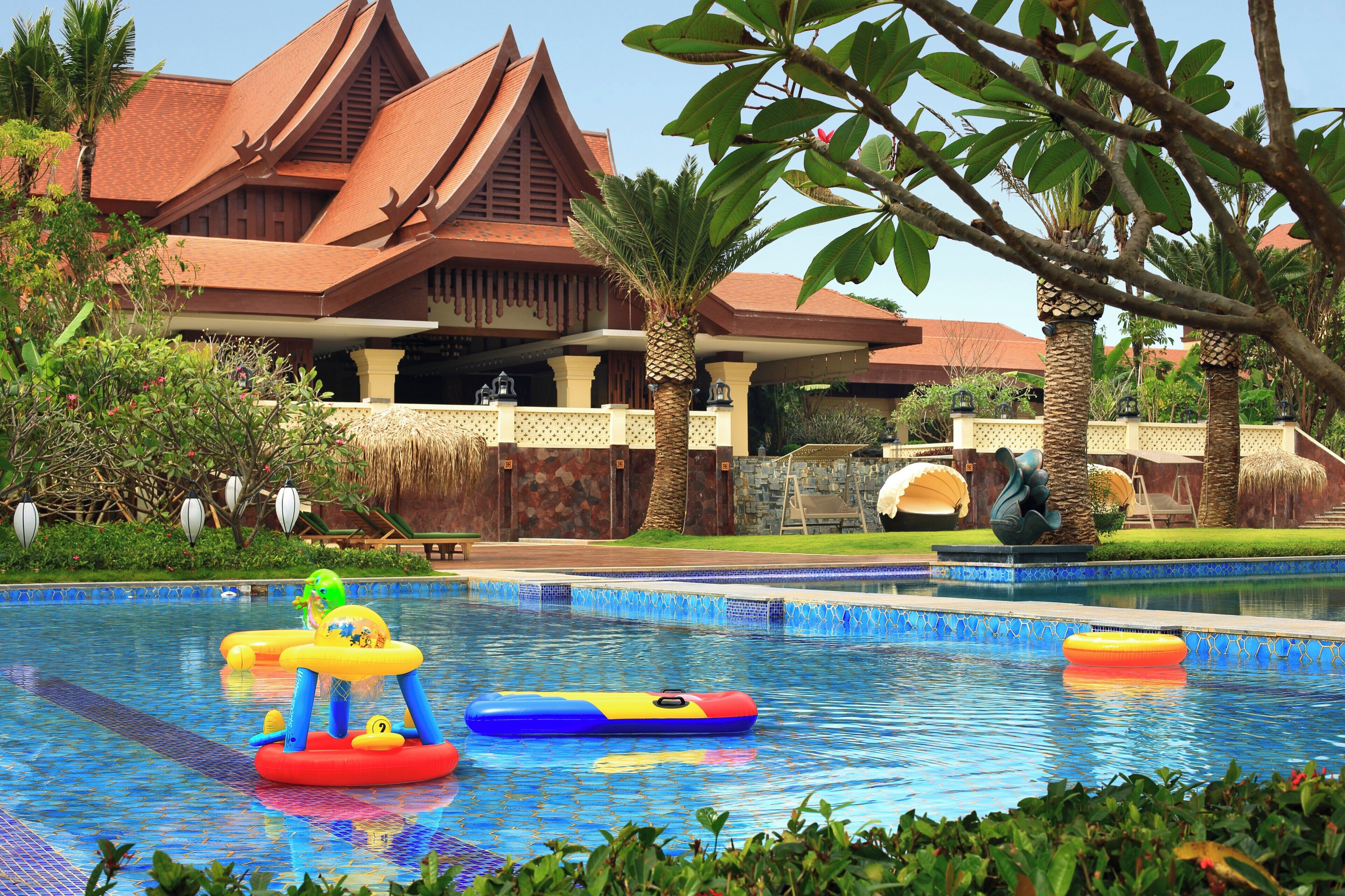 A swimming pool with inflatable toys, palm trees, and a traditional building with a red roof.