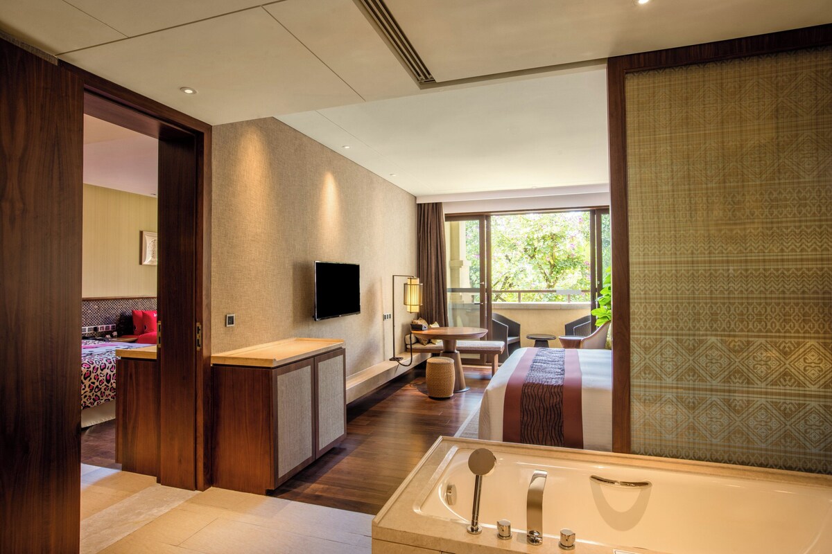 Quarto de hotel com uma banheira grande, sofá, TV e varanda com vista para o verde.