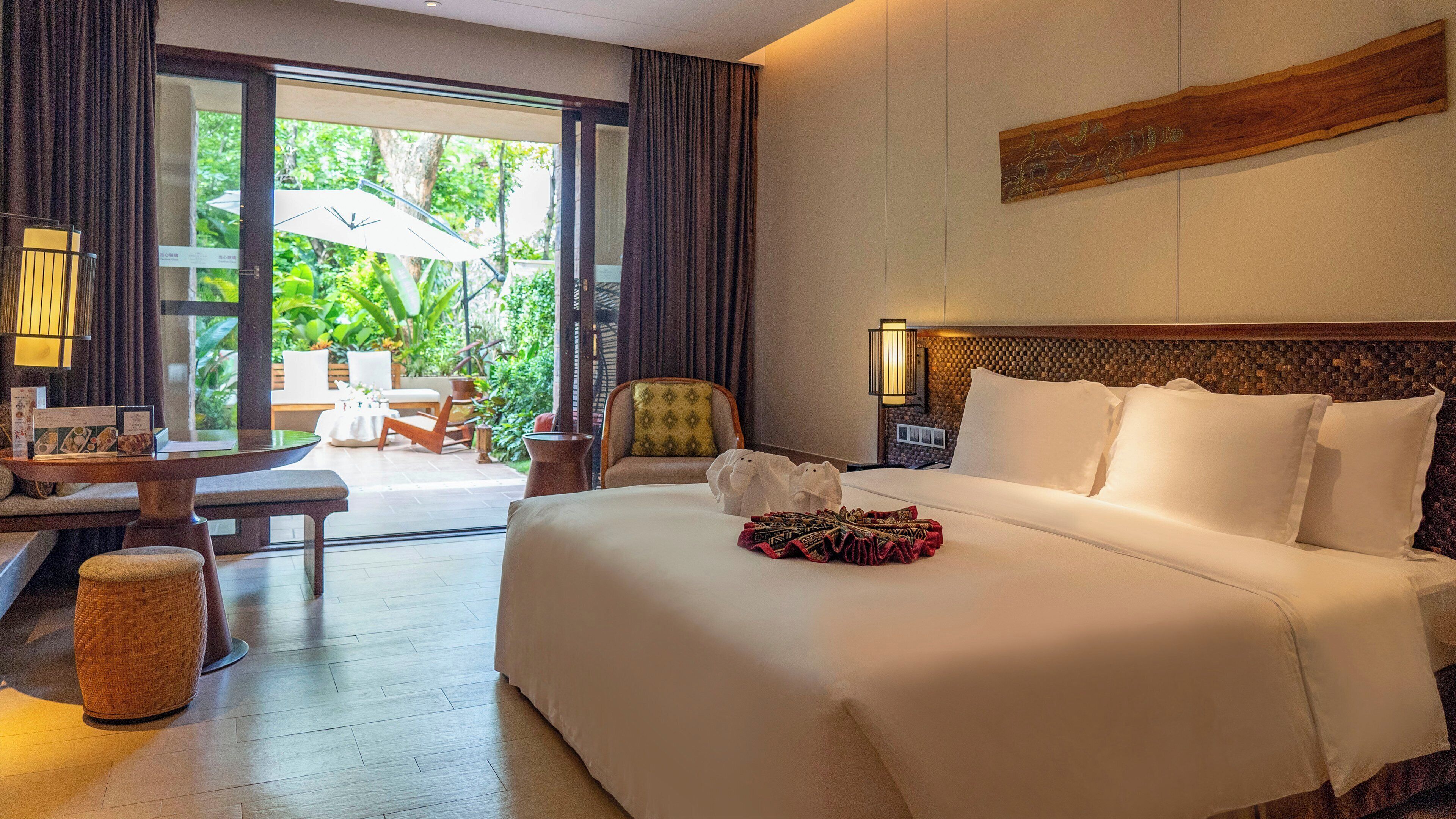 A hotel room with a large bed, a desk with a lamp, a chair, and a view of greenery outside.
