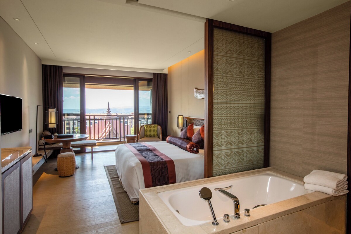 Quarto de hotel com varanda, uma cama grande, escrivaninha, poltrona, televisão e banheira.