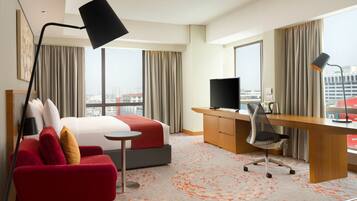 A hotel room with a bed, a desk, a chair, a TV, and large windows offering a city view.