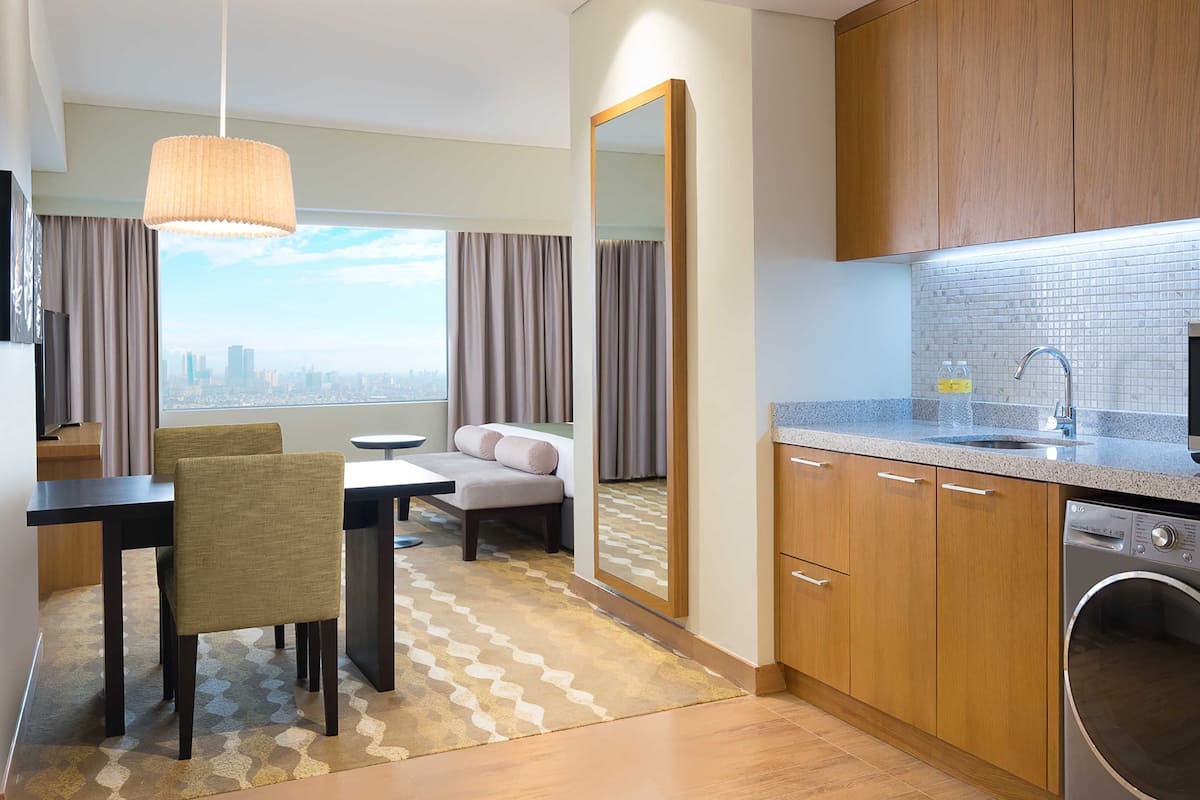 Hotelkamer met een eethoek, een bank, een groot raam met uitzicht over de stad, en een keuken met een magnetron, oven en wasmachine.