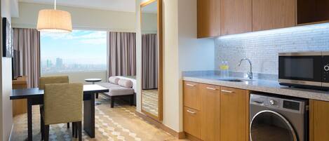 Habitación de hotel con comedor, sofá, ventana grande con vista a la ciudad, cocina equipada con microondas, horno y lavadora.