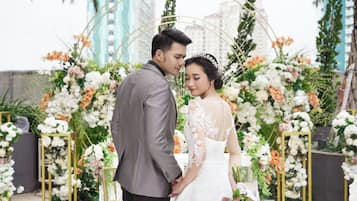 A couple holding hands in front of a floral arch with a cityscape in the background.