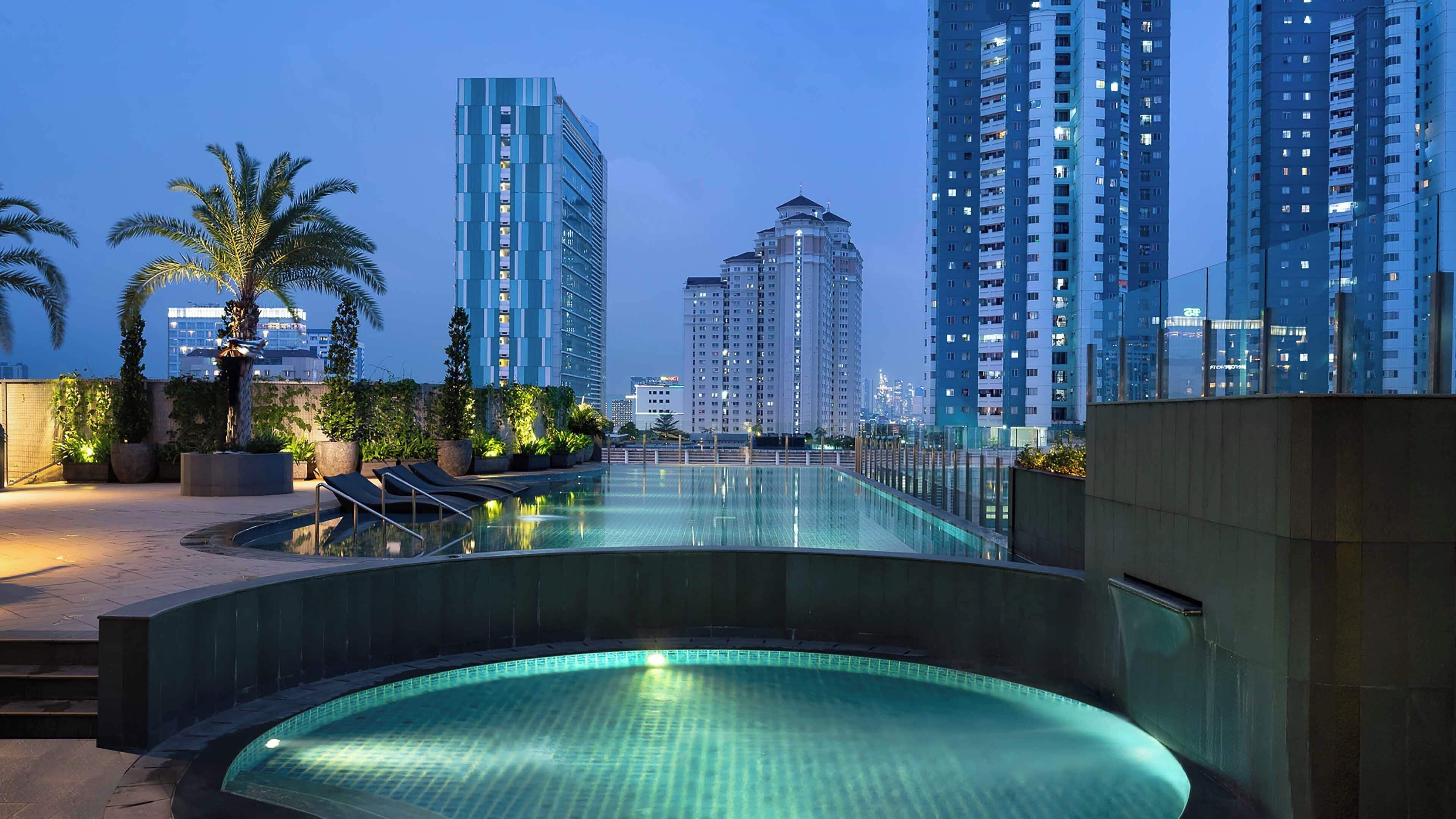 Una piscina en la azotea con sillones de descanso, rodeada de edificios altos y palmeras.