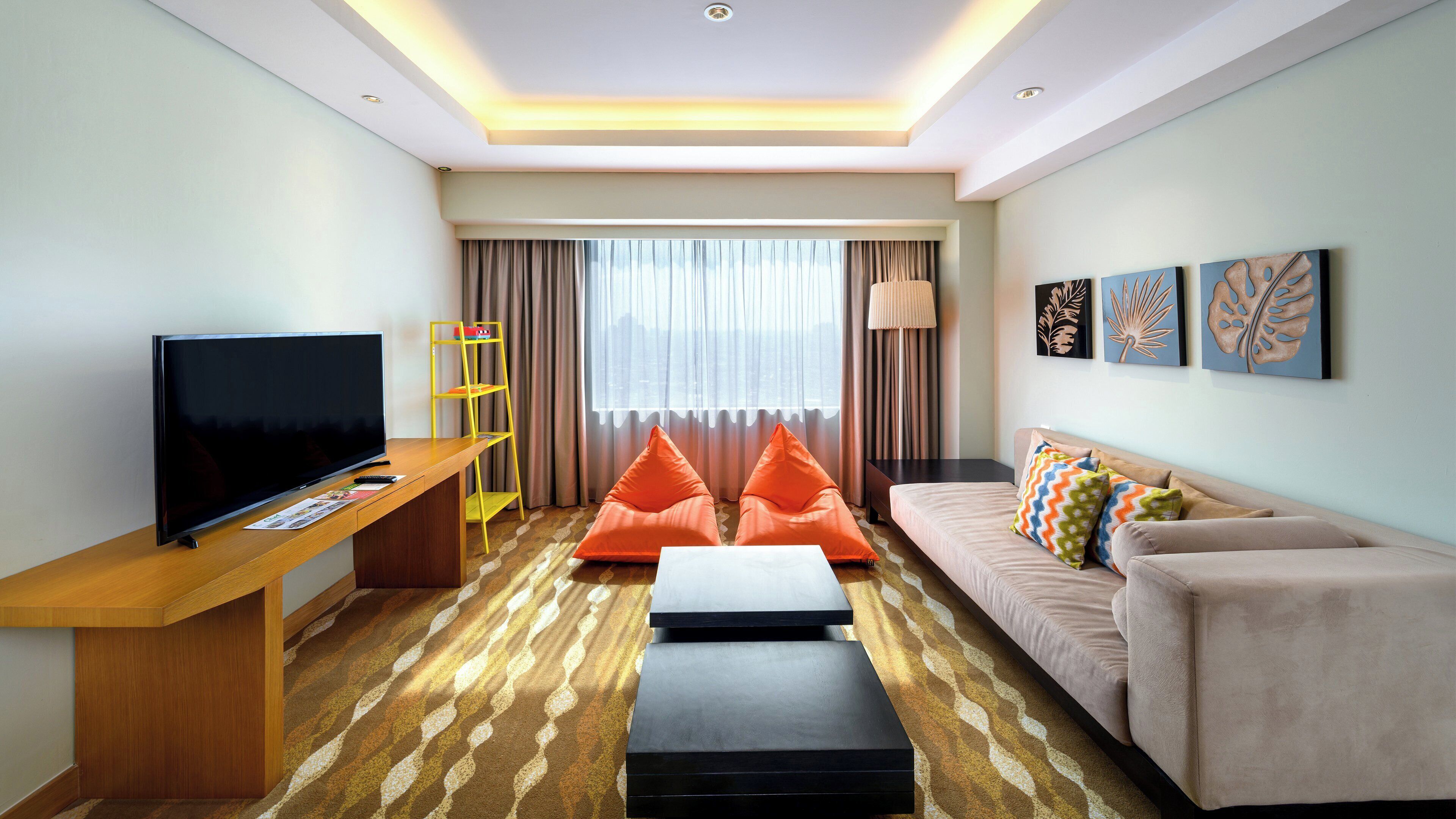 Ein modernes Hotelzimmer mit Fernseher, Schreibtisch aus Holz, einer Couch, orangenen Sitzsäcken, einem gemusterten Teppich und Bildern an den Wänden.