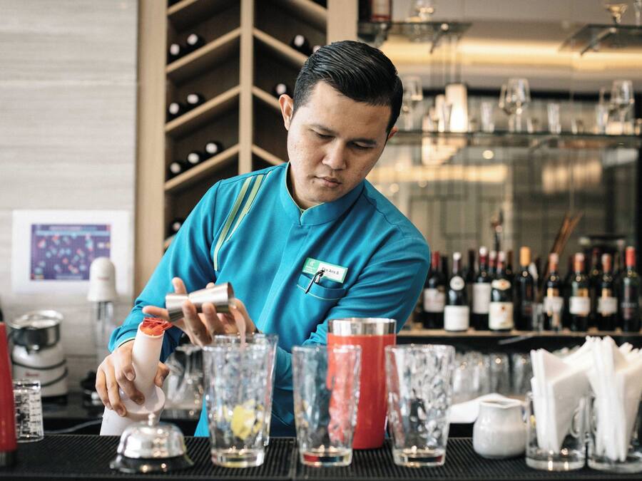 A bartender preparing drinks at a bar with a wine rack and various bottles in the background.