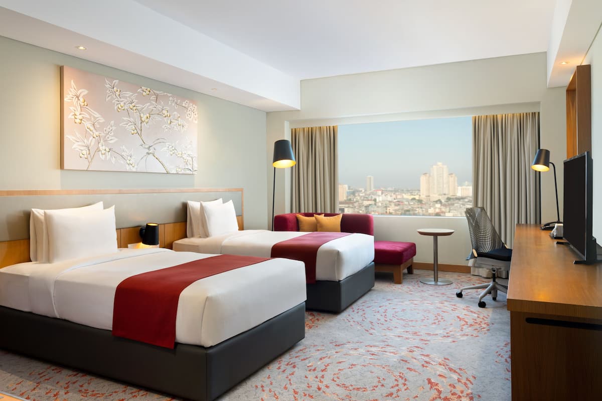 Een hotelkamer met twee bedden, een bureau, een stoel en een groot raam met uitzicht op de stad.