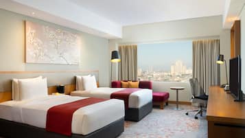 Habitación de hotel con dos camas, un escritorio, una silla y un ventanal con vista a la ciudad.