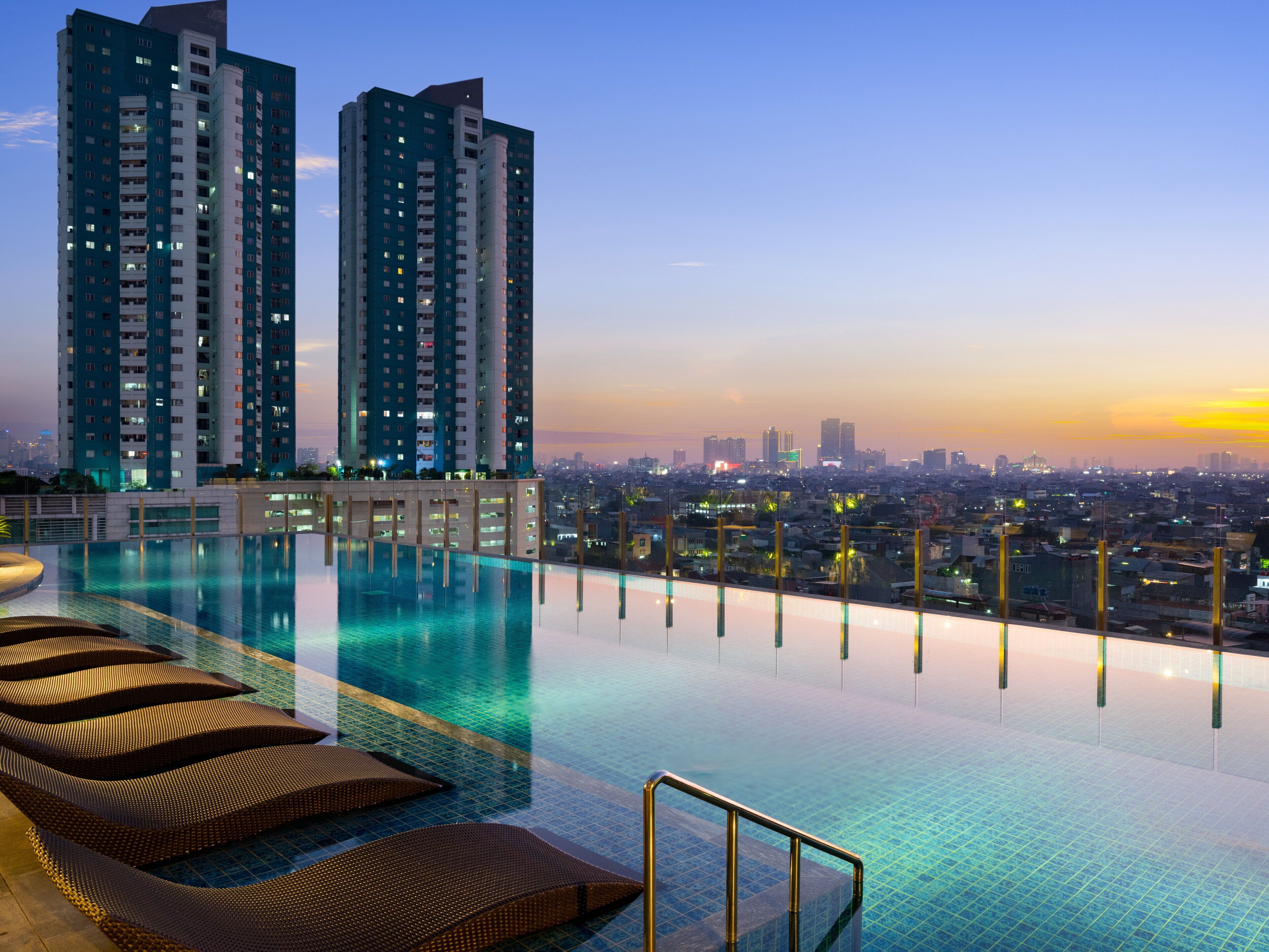 A rooftop infinity pool with lounge chairs, surrounded by high-rise buildings and a cityscape at sunset.