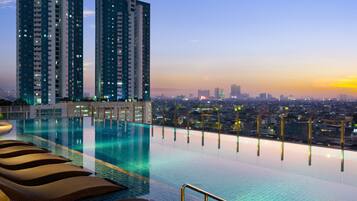 A rooftop infinity pool with lounge chairs, surrounded by high-rise buildings and a cityscape at sunset.