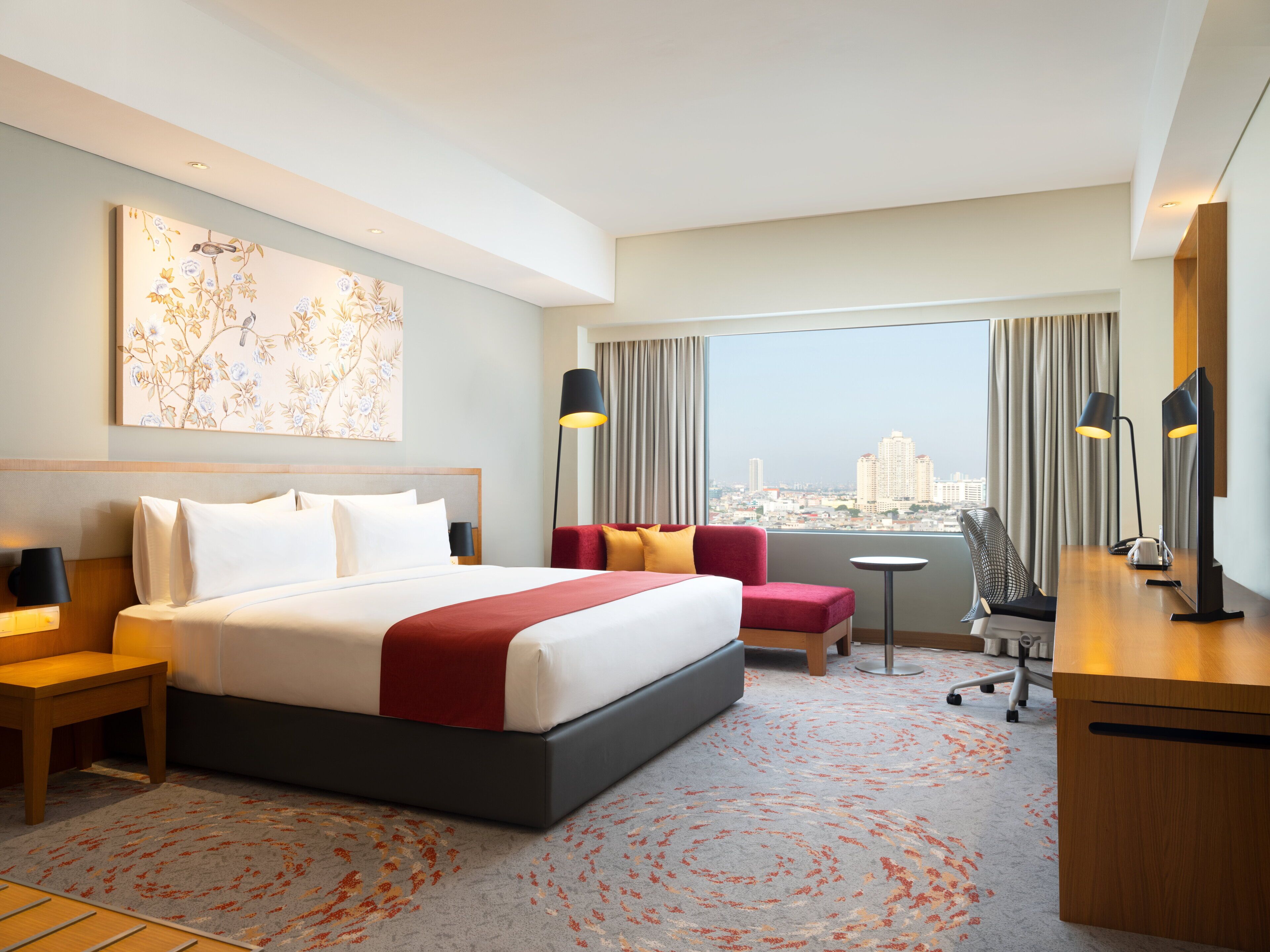 A hotel room with a bed, a desk, a chair, a small table, and a large window with a city view.
