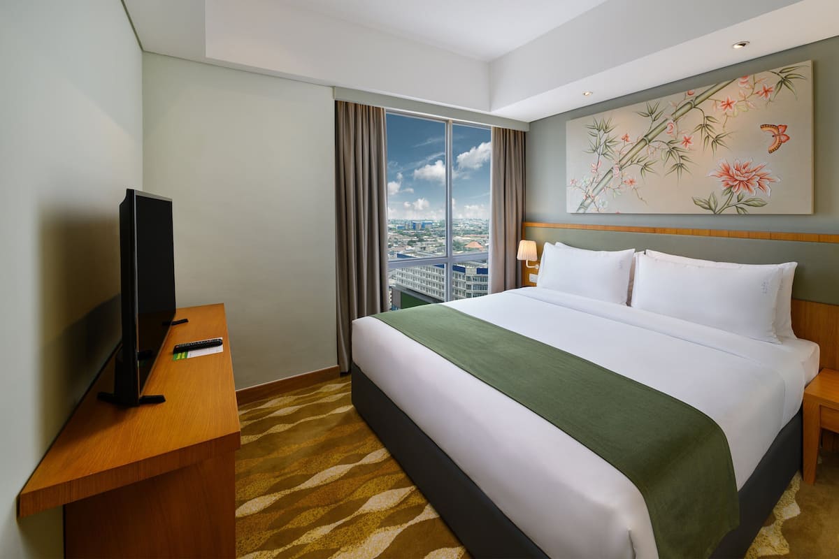 Hotelkamer met een bed, een televisie op een houten bureau, een nachtkastje met een lamp en uitzicht op de stad door grote ramen.