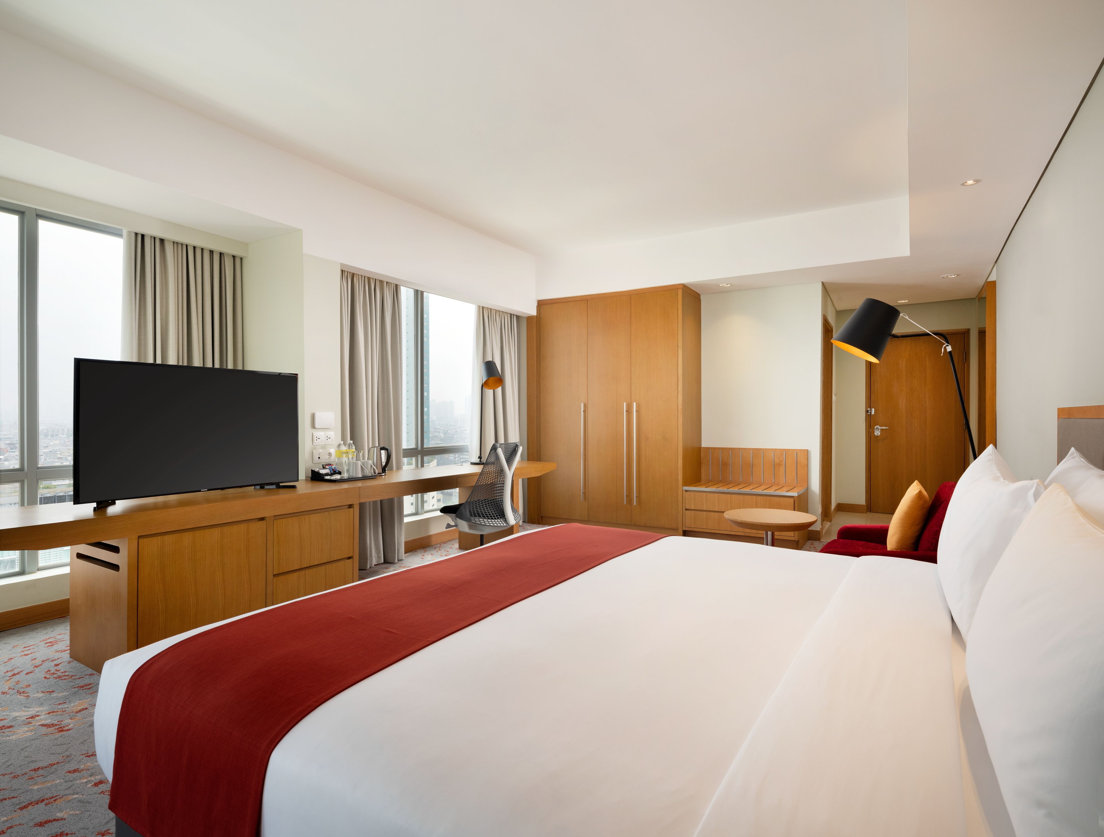 A modern hotel room with a large bed, a desk with a flat-screen TV, a chair, a desk lamp, and a wardrobe.