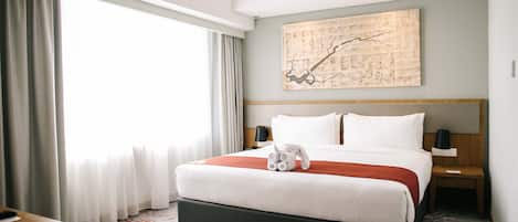 Habitación de hotel con una cama grande, una mesita de noche, una lámpara, un cuadro en la pared y vistas al exterior a través de la ventana.