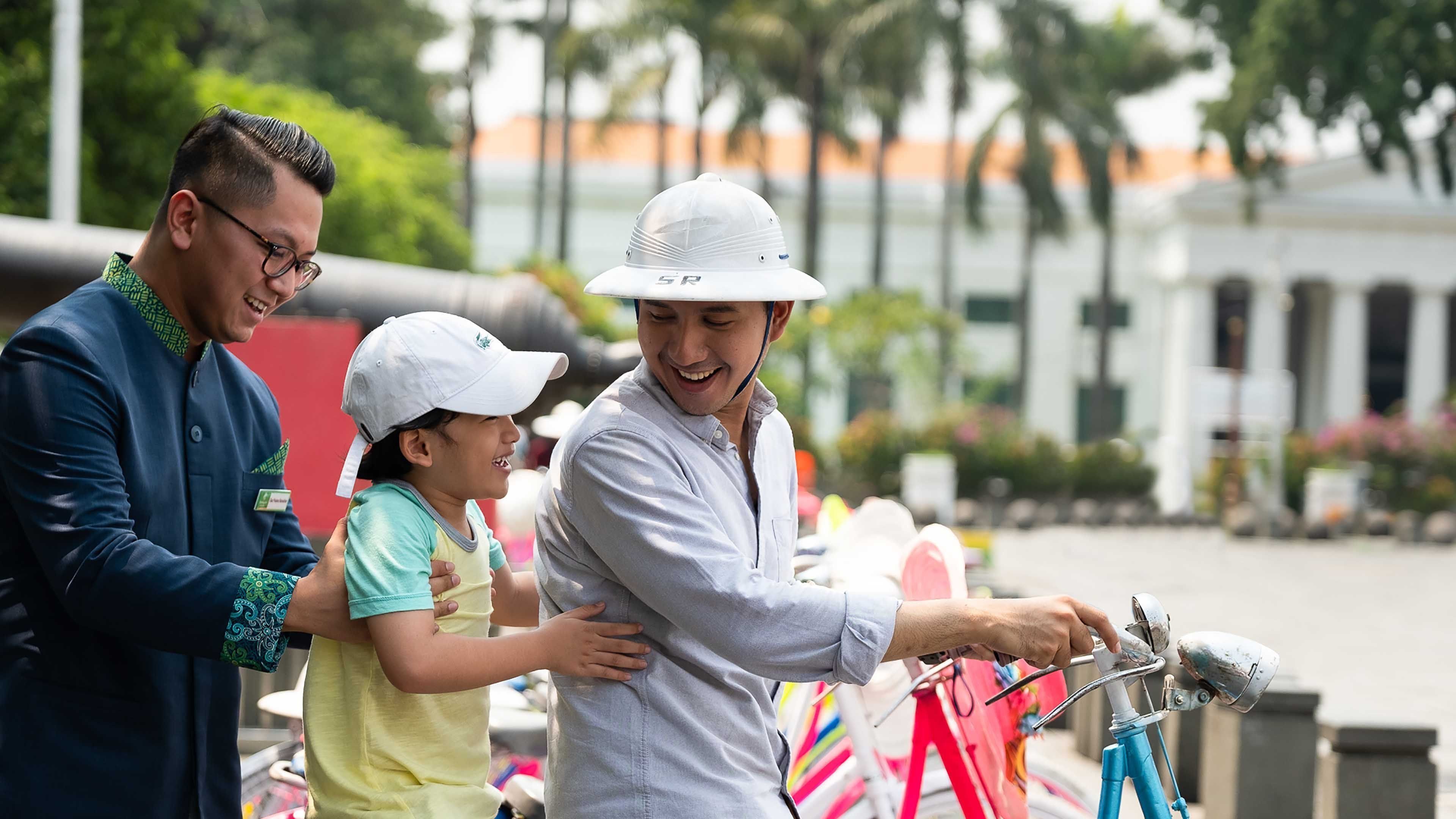 A man in a white hat and light blue shirt working on a bicycle with a child in a yellow shirt and white cap nearby.