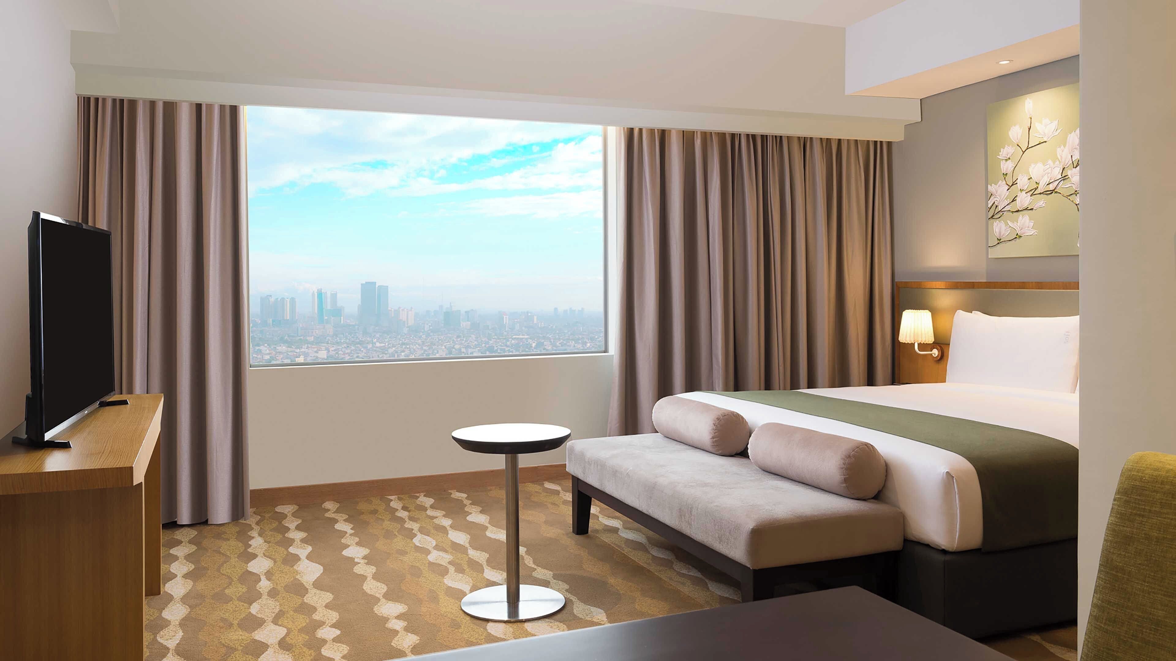 A hotel room with a bed, a TV, a small table, and a view of a cityscape.