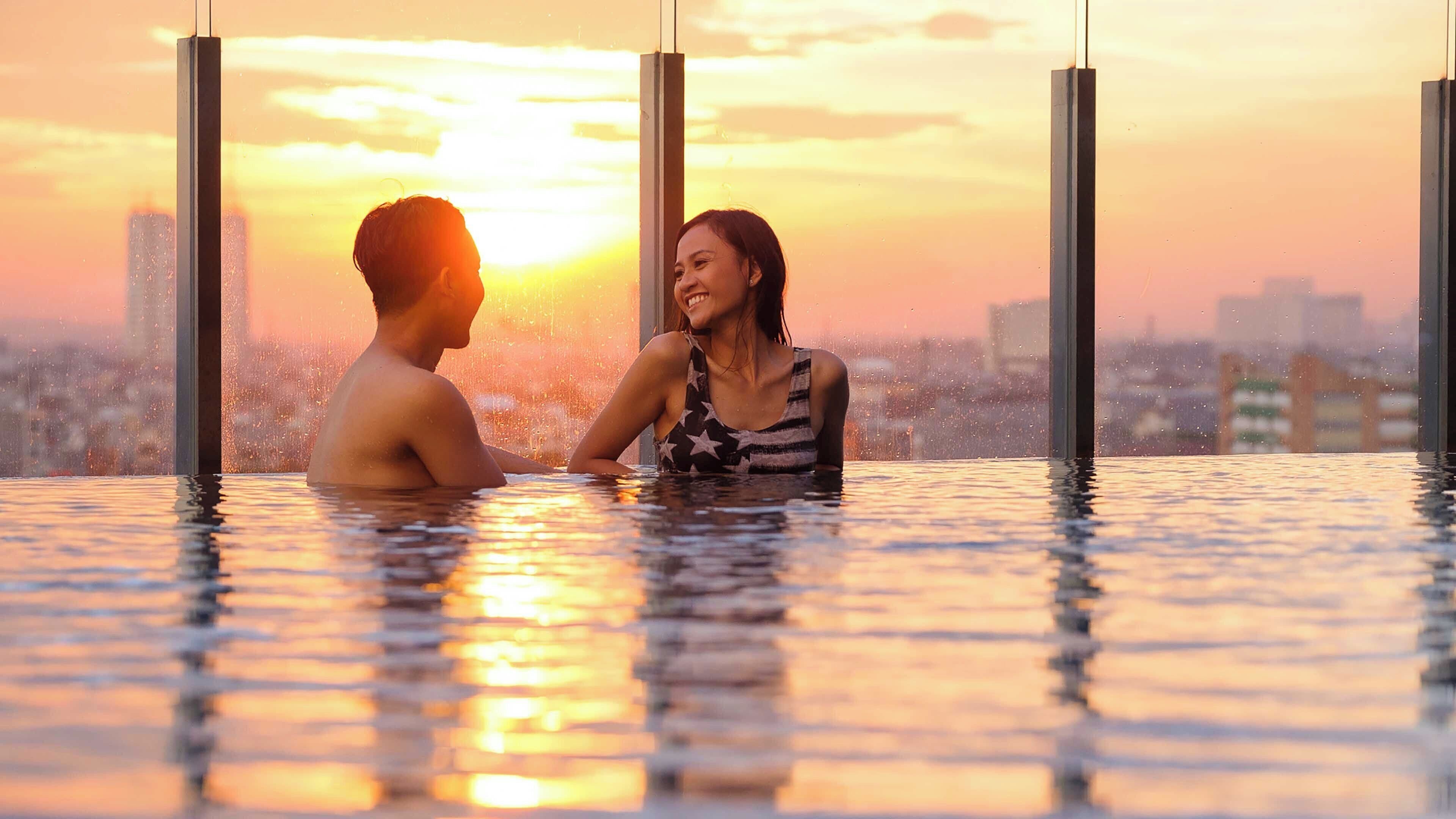 Una pareja en una piscina exterior climatizada, con un paisaje urbano al fondo.
