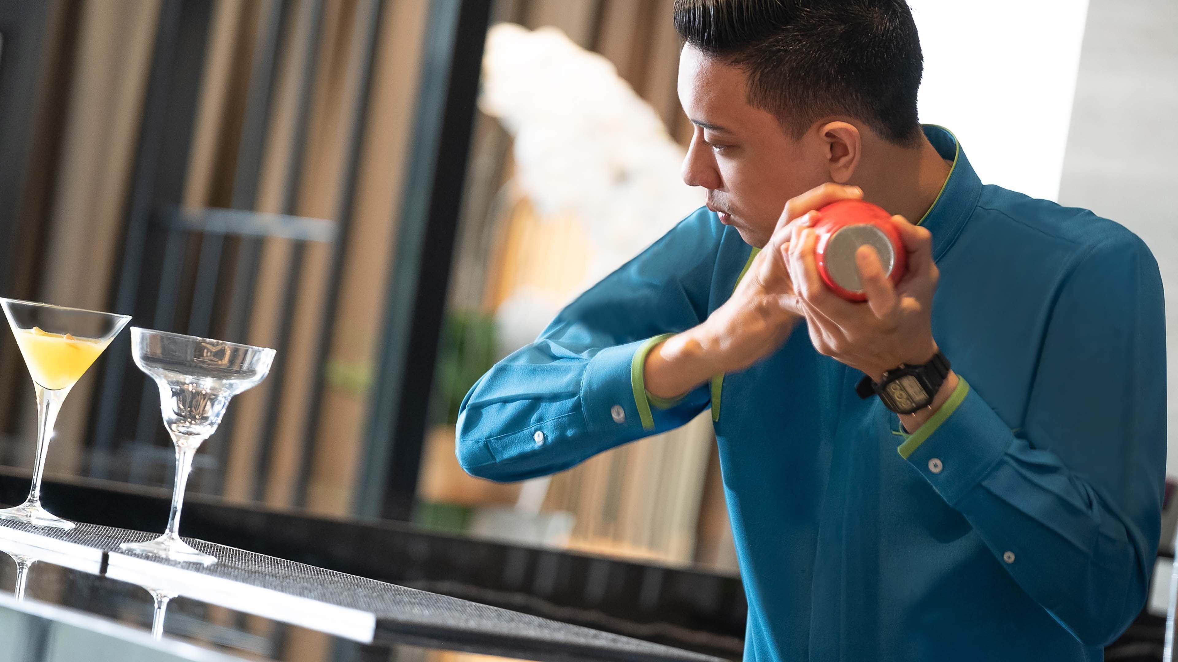 A person in a blue shirt preparing a drink with a shaker. Two martini glasses are on the counter.