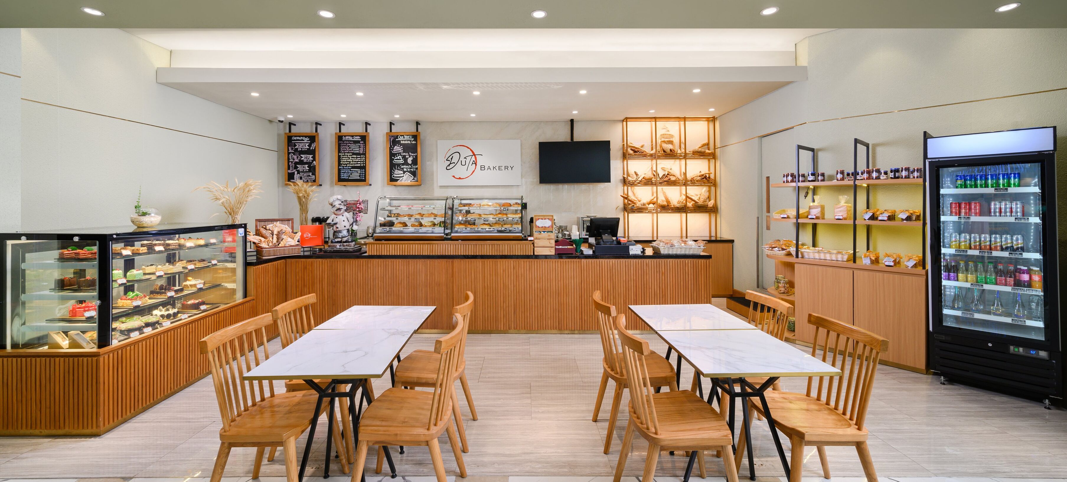 Una panadería con mesas y sillas de madera, un expositor de productos horneados y un dispensador de bebidas refrigeradas.