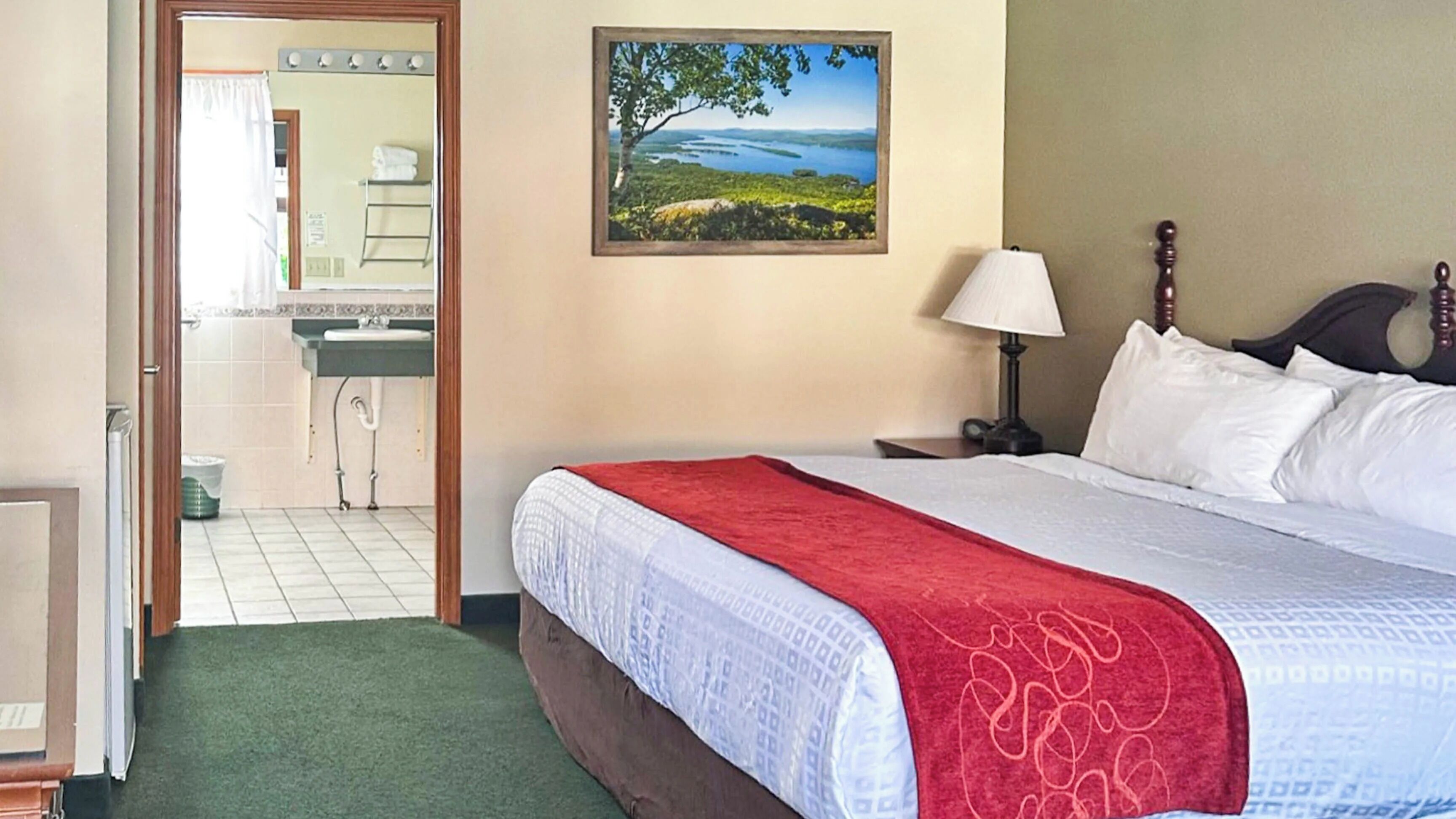 A hotel room with a bed, a nightstand, a lamp, a painting, and a bathroom visible through an open door.