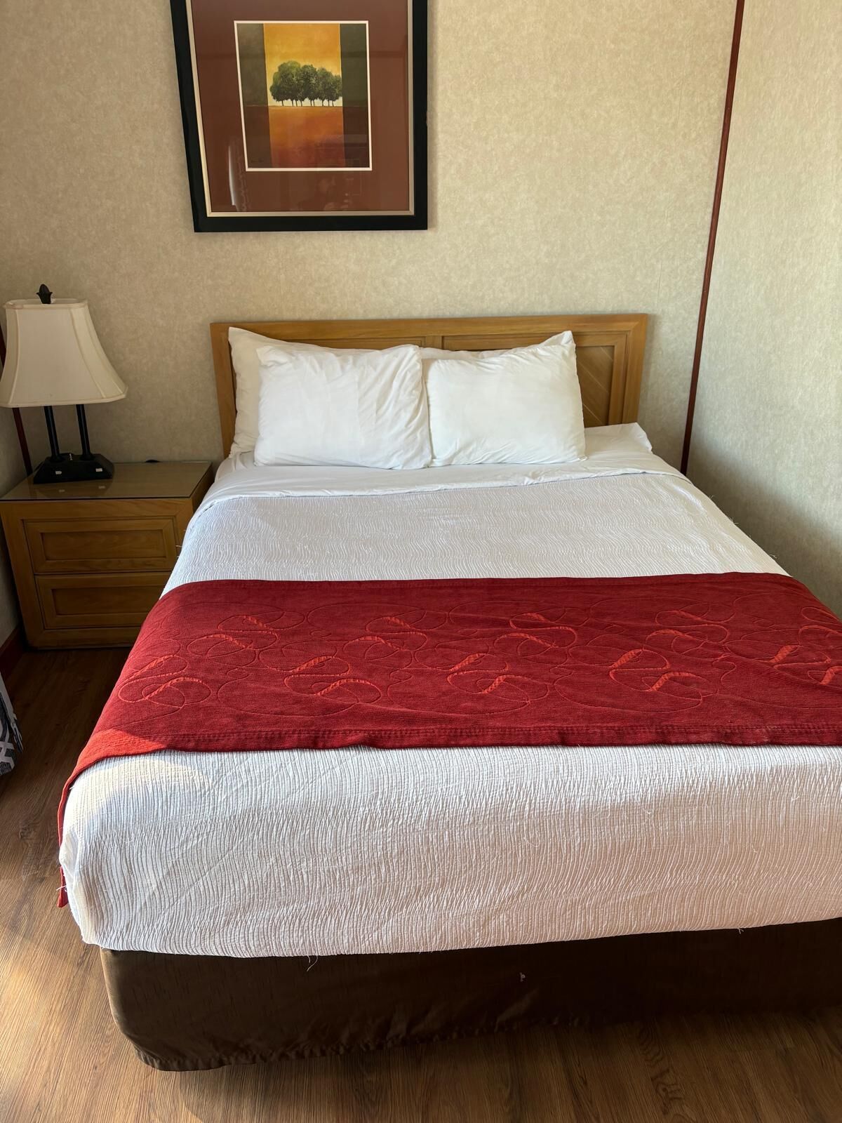 Una cama bien tendida con sábanas blancas y una manta roja, un cabecero de madera, una mesita de noche con una lámpara y un cuadro en la pared.