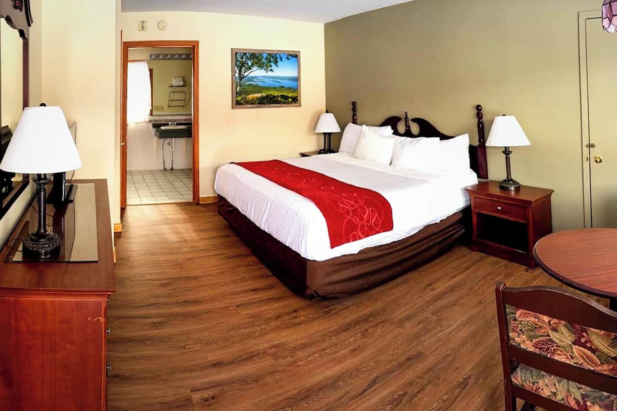 A hotel room with a bed, two bedside tables with lamps, a wooden chair, a small round table, a painting on the wall, and a door leading to a bathroom.