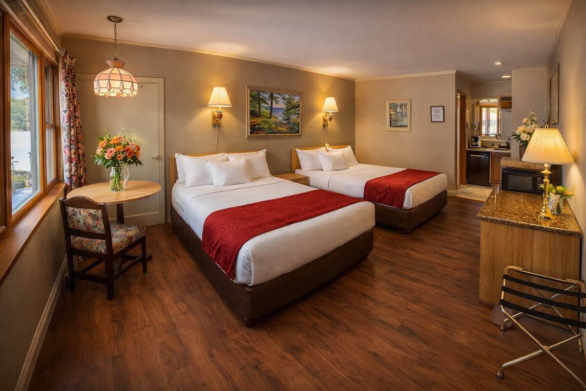 A hotel room with two beds, a dining table, a small kitchen area, and a view of the outdoors.