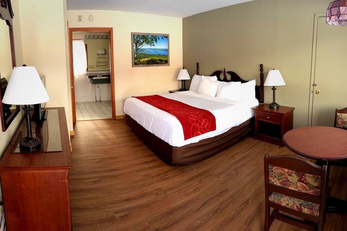 A hotel room with a bed, a desk, a chair, a round table, a mirror, a picture on the wall, and a bathroom visible through an open door.