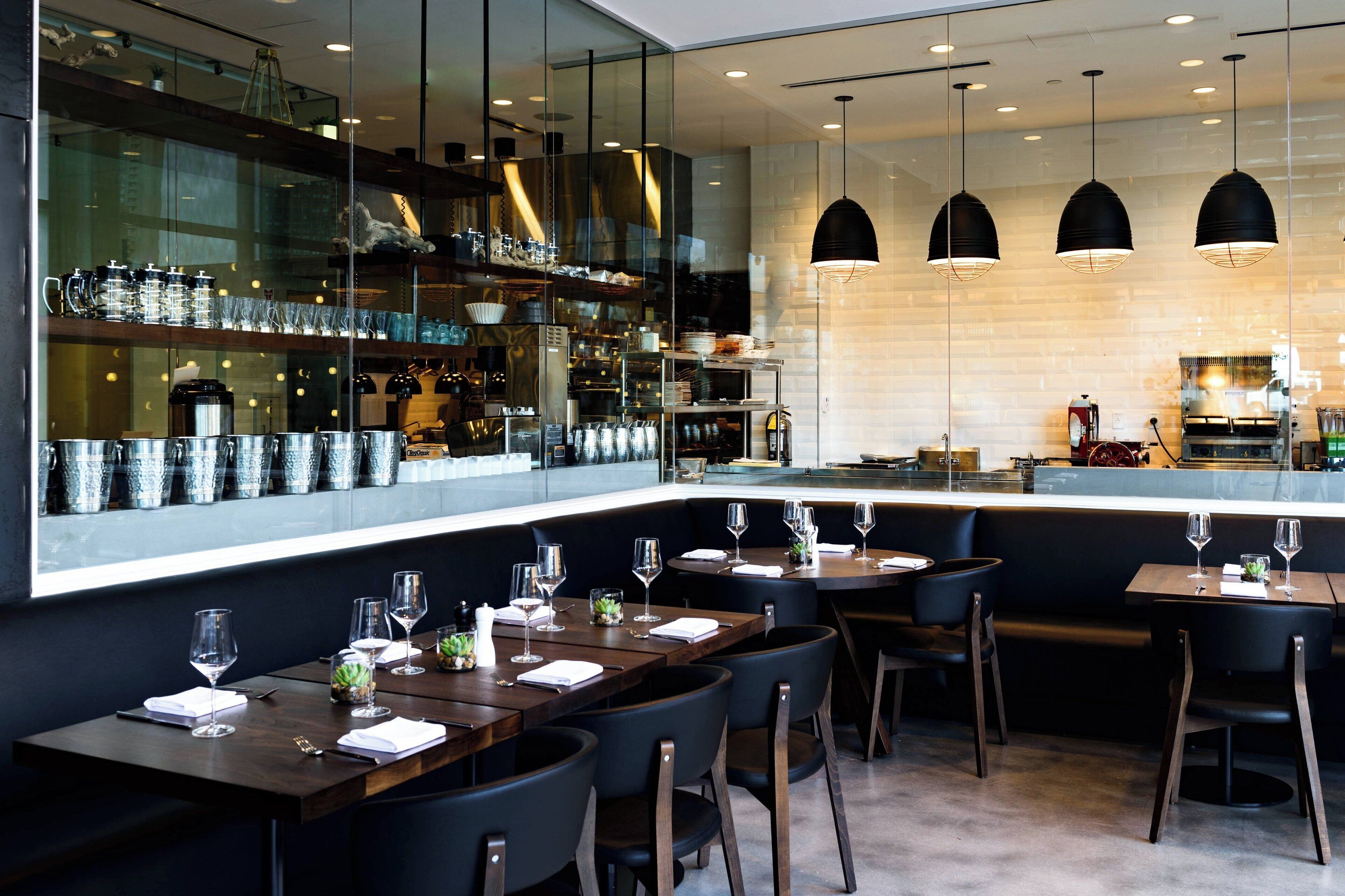 A modern restaurant with dark wooden tables and chairs, a glass wall displaying kitchen items, and pendant lighting.