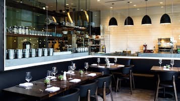 A modern restaurant with dark wooden tables and chairs, a glass wall displaying kitchen items, and pendant lighting.