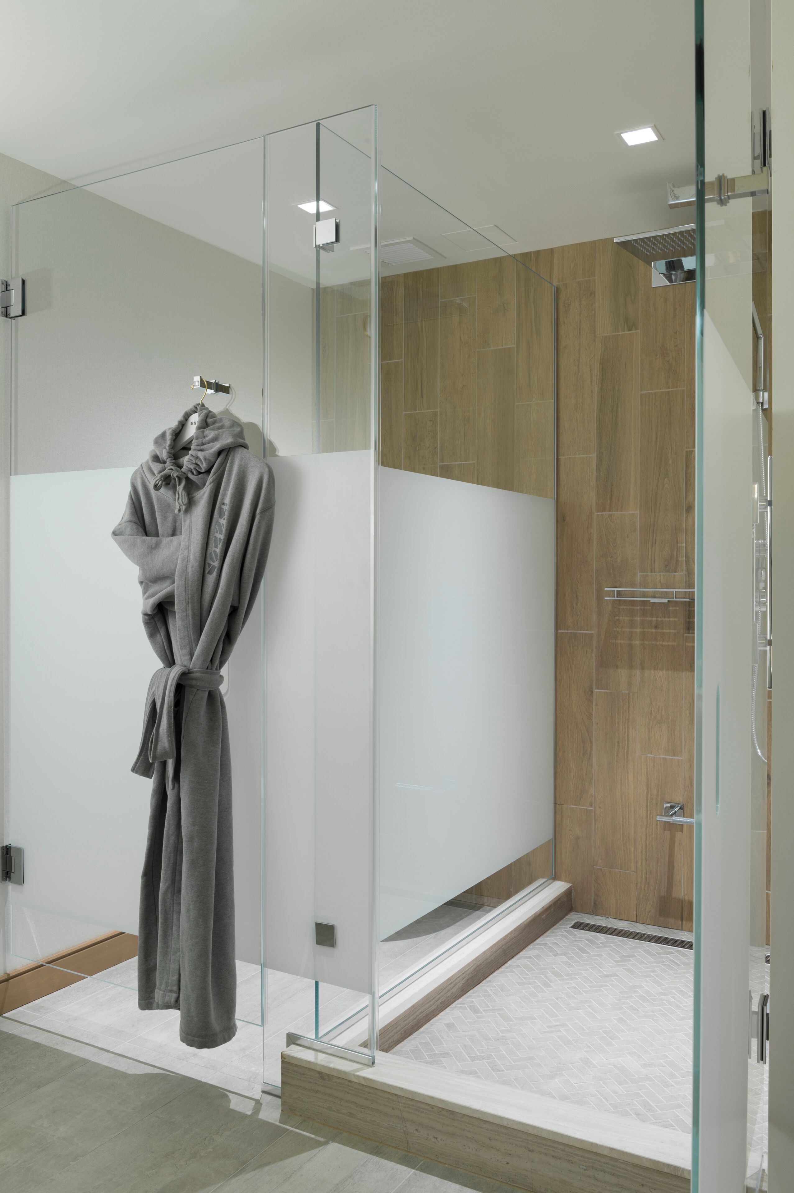 Un baño moderno con cabina de ducha de vidrio, suelo de madera y una bata gris colgada en la pared.