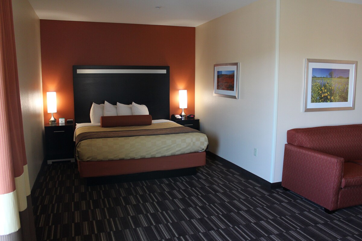 Un cuarto de hotel con una cama grande, dos mesitas de noche con lámparas, un sillón rojo y dos cuadros en la pared.
