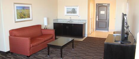 A hotel room with a red sofa, a small table, a kitchen area with a sink, and a door leading to another room.