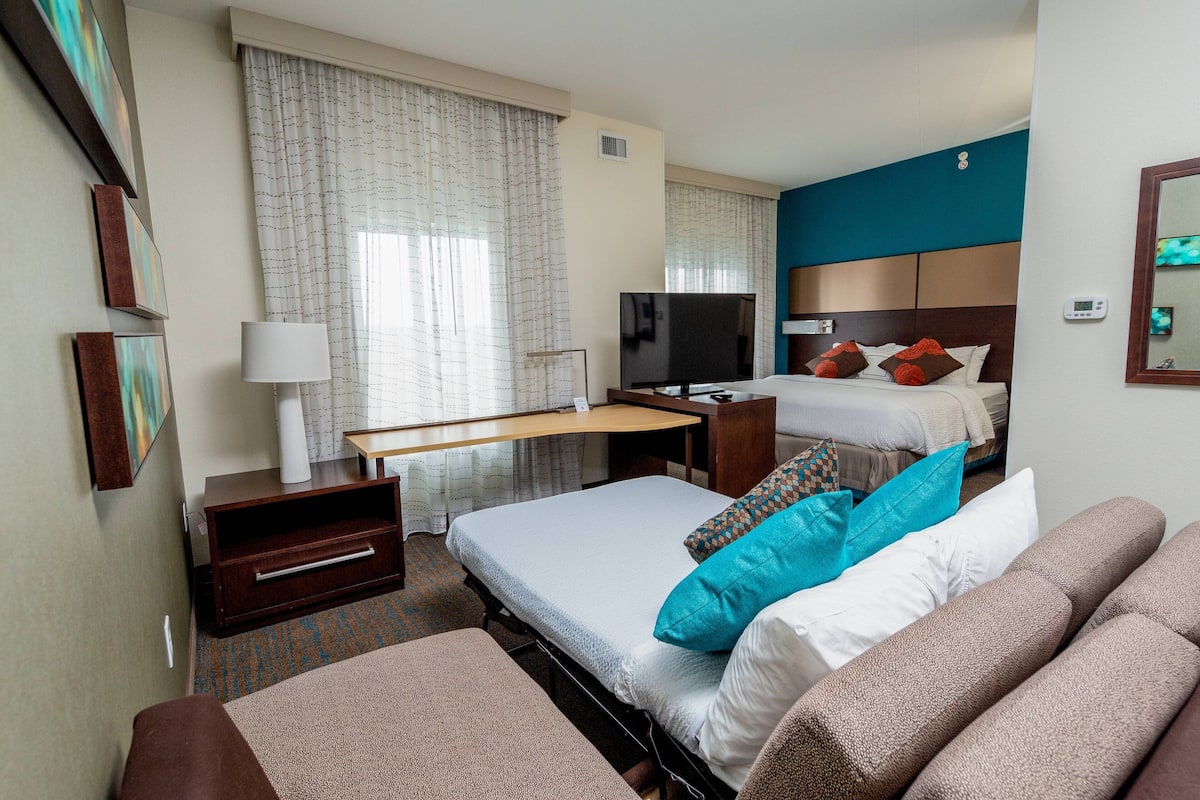 Habitación de hotel con cama, escritorio, televisión y un amplio ventanal con cortinas.