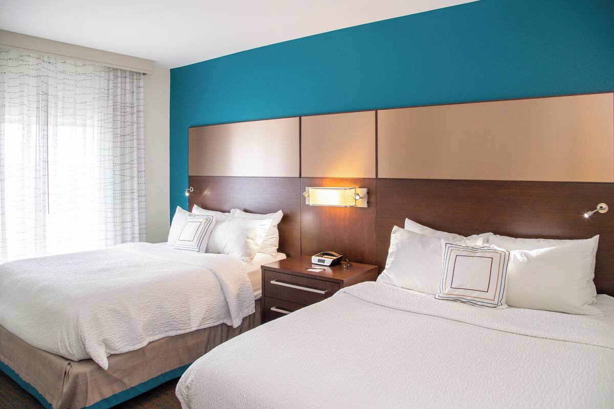 A hotel room with two beds, a nightstand with a lamp, and a window with curtains.
