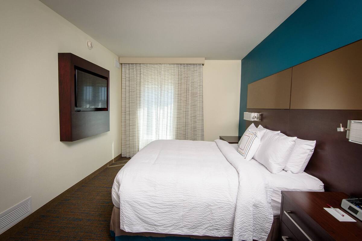 A hotel room with a large bed, a flat-screen TV mounted on the wall, a bedside table with a lamp, and a window with curtains.